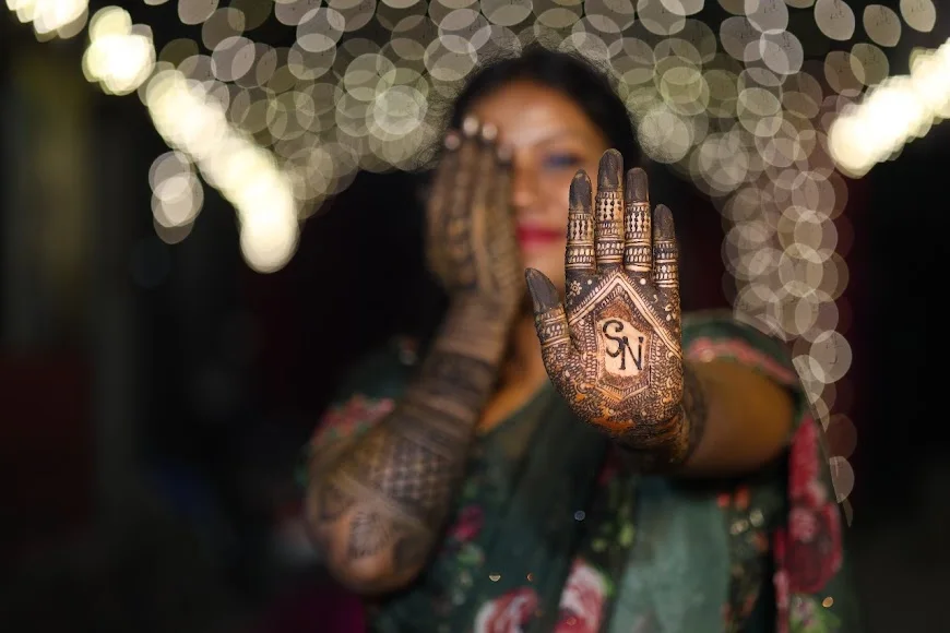 A woman displaying intricate mehndi on her raised hand with warm lights behind her